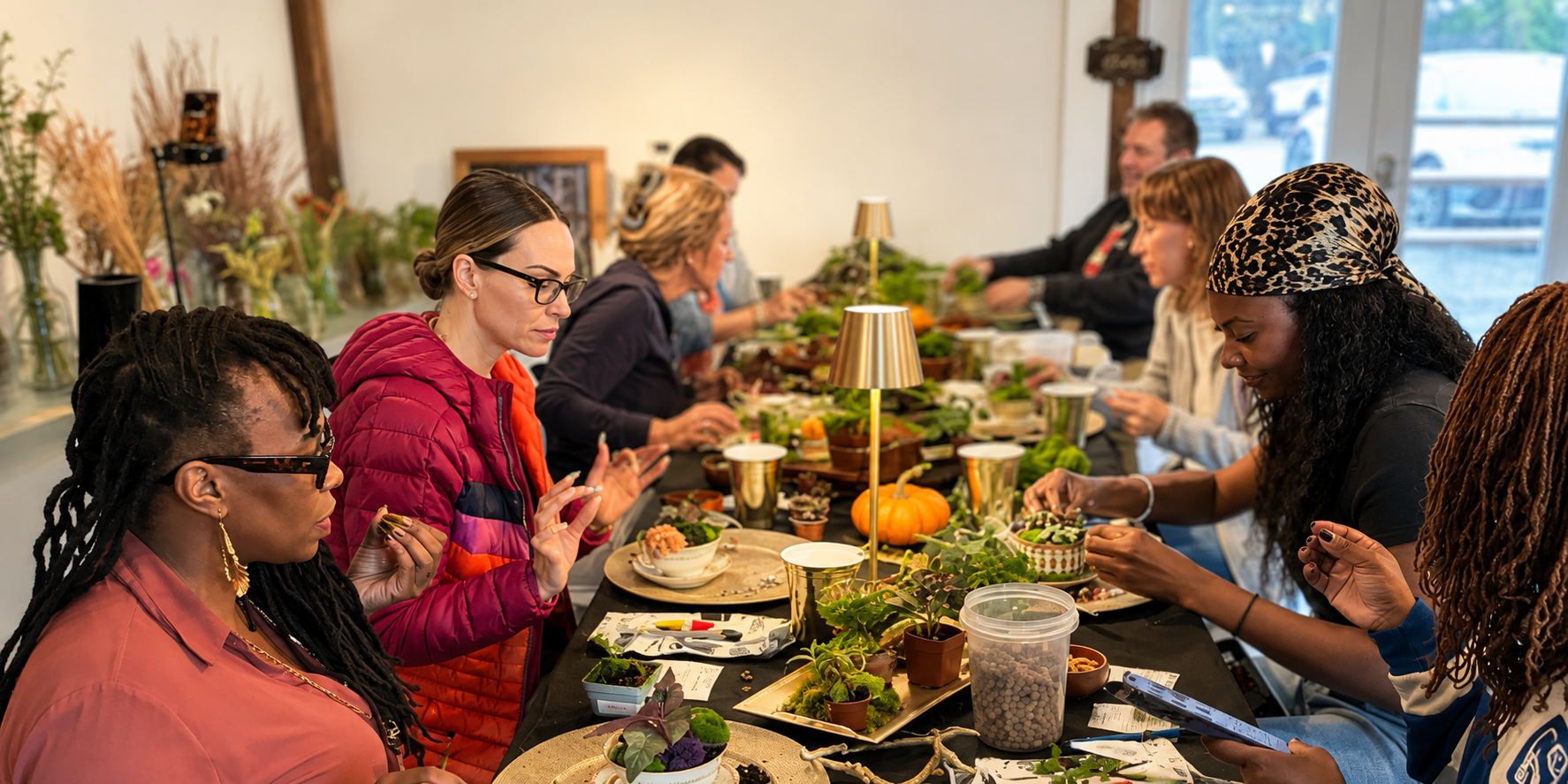 A group of people sitting around a long table at a plant workshop, working with small plants and moss under warm lamps